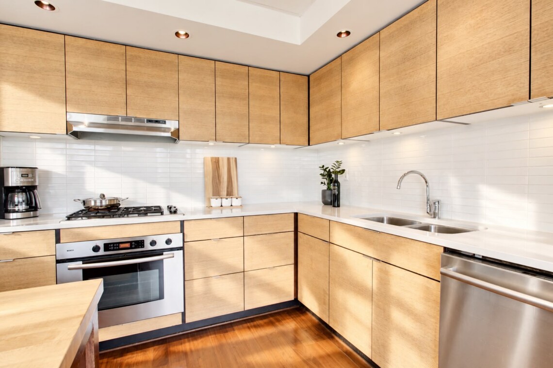 Modern L-shaped kitchen with light wood cabinets, white tiled backsplash, and stainless steel appliances including oven, stove, and dishwasher.
