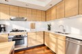 Modern L-shaped kitchen with light wood cabinets, white tiled backsplash, and stainless steel appliances including oven, stove, and dishwasher.