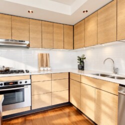 Modern L-shaped kitchen with light wood cabinets, white tiled backsplash, and stainless steel appliances including oven, stove, and dishwasher.