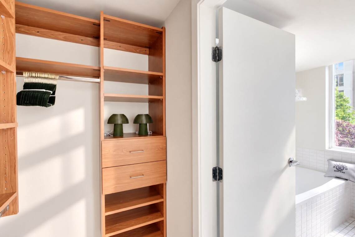 Wooden closet with open shelving, a hanging rod with folded clothes, and a two-drawer cabinet; two green lamps on a shelf and sunlight casting shadows; doorway leads to a bright tiled bathroom area visible through the opening.