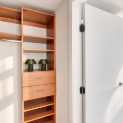 Wooden closet with open shelving, a hanging rod with folded clothes, and a two-drawer cabinet; two green lamps on a shelf and sunlight casting shadows; doorway leads to a bright tiled bathroom area visible through the opening.