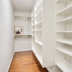 A small walk-in closet with white shelves and a wooden floor; a framed black-and-white photo sits on a clothing rod at the back wall.