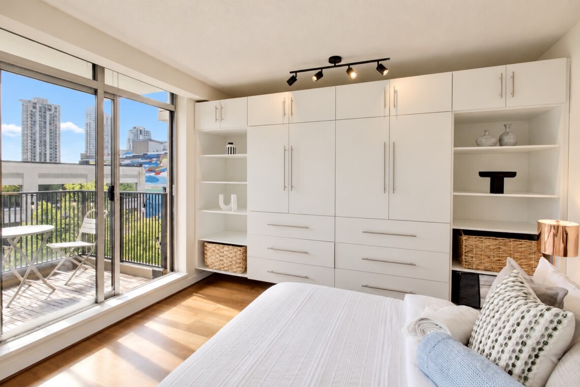 Bright bedroom with a wall of white built-in wardrobes and open shelves flanking a central storage unit, overlooking a balcony with city buildings outside. A bed with white linens is in the foreground and track lighting highlights the space.