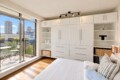 Bright bedroom with a wall of white built-in wardrobes and open shelves flanking a central storage unit, overlooking a balcony with city buildings outside. A bed with white linens is in the foreground and track lighting highlights the space.