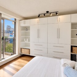 Bright bedroom with a wall of white built-in wardrobes and open shelves flanking a central storage unit, overlooking a balcony with city buildings outside. A bed with white linens is in the foreground and track lighting highlights the space.