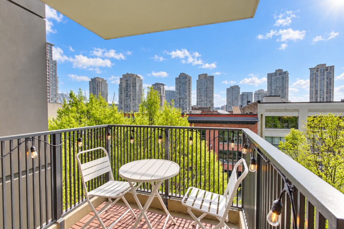 A small balcony with white metal table and two matching chairs, string lights along a black railing, overlooking a city skyline and green trees under a blue sky.