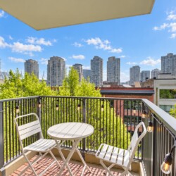 A small balcony with white metal table and two matching chairs, string lights along a black railing, overlooking a city skyline and green trees under a blue sky.