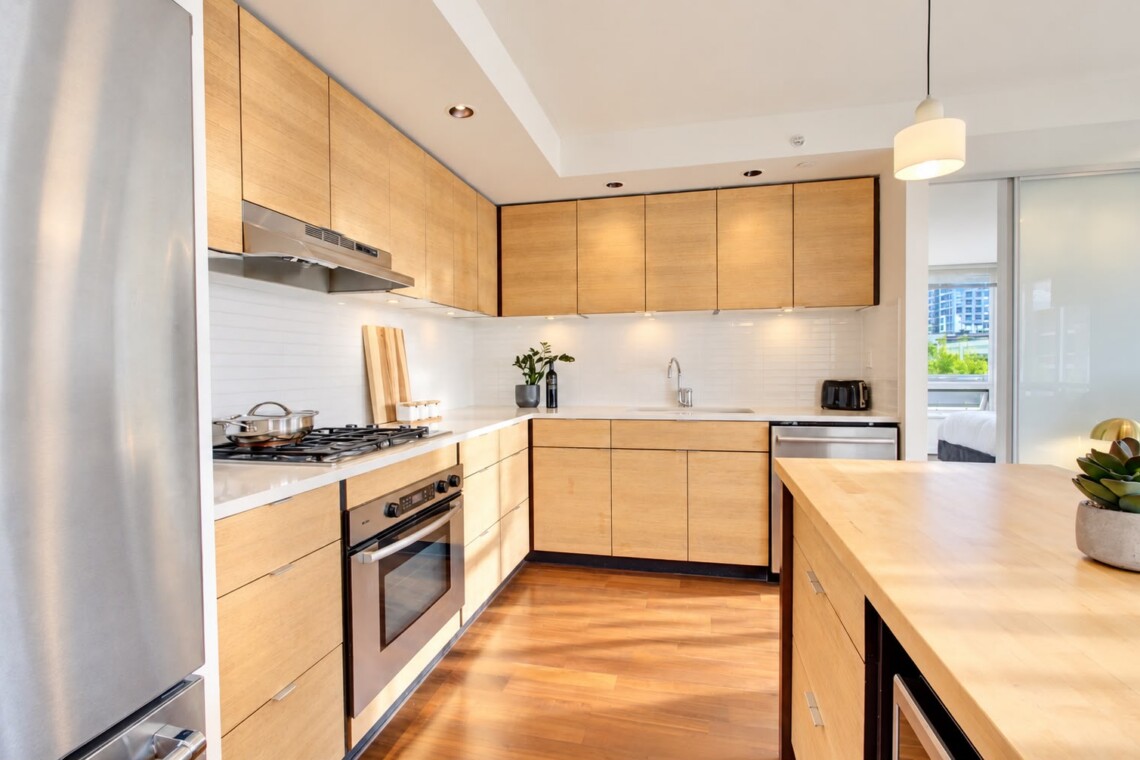Modern L-shaped kitchen with light wood cabinets, stainless appliances, and a large central island.