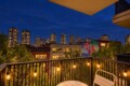 Balcony at dusk with warm string lights, a small table and chair, overlooking trees and a city skyline with a colorful mural on a nearby wall.