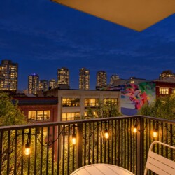 Balcony at dusk with warm string lights, a small table and chair, overlooking trees and a city skyline with a colorful mural on a nearby wall.