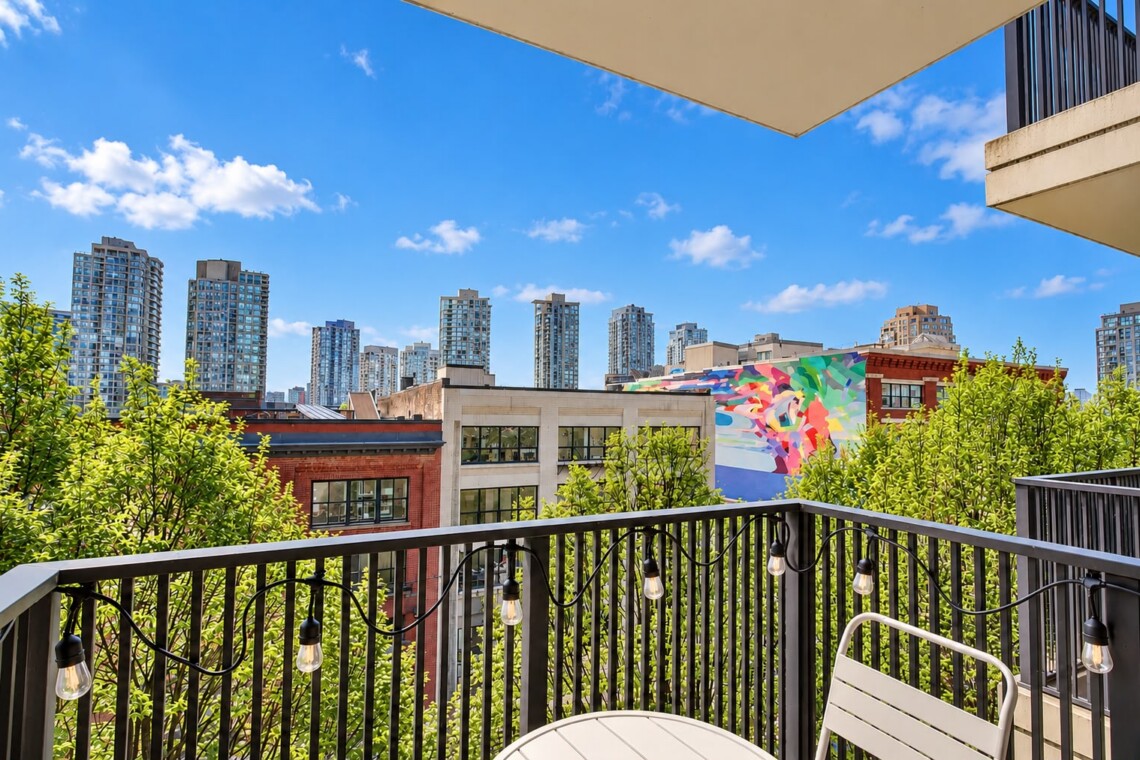 Balcony view of a city with a colorful mural, green trees, and string lights along a black railing.