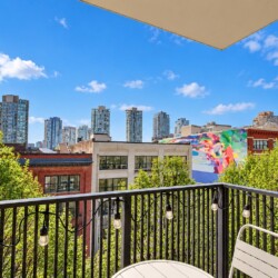Balcony view of a city with a colorful mural, green trees, and string lights along a black railing.