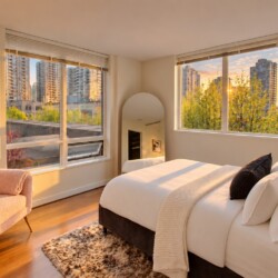 Sunlit modern bedroom with a white bed and pillows, a pink upholstered chair, and a large arched mirror by the window overlooking a cityscape.