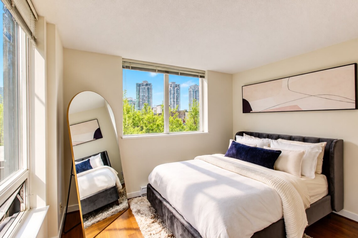 Bright bedroom with a white bed, dark headboard, abstract wall art, and a large window showing a city skyline outside.