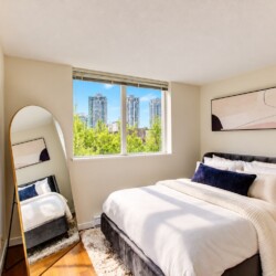Bright bedroom with a white bed, dark headboard, abstract wall art, and a large window showing a city skyline outside.