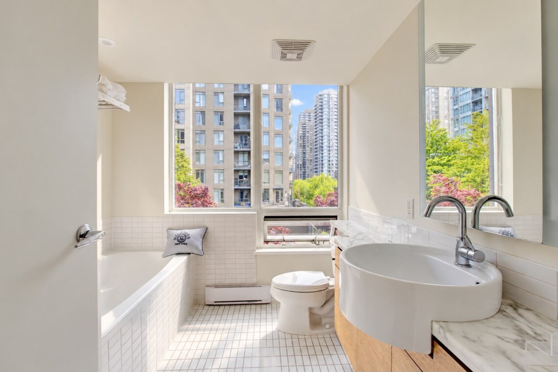 Bright urban bathroom with a white tiled tub, toilet, and sleek curved sink, cool gray walls, and a large window showing city buildings and trees outside.