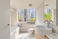 Bright urban bathroom with a white tiled tub, toilet, and sleek curved sink, cool gray walls, and a large window showing city buildings and trees outside.