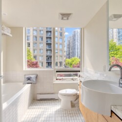 Bright urban bathroom with a white tiled tub, toilet, and sleek curved sink, cool gray walls, and a large window showing city buildings and trees outside.