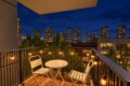 Nighttime balcony with string lights, a white metal table and two chairs, overlooking a city skyline.