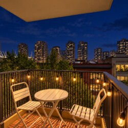 Nighttime balcony with string lights, a white metal table and two chairs, overlooking a city skyline.