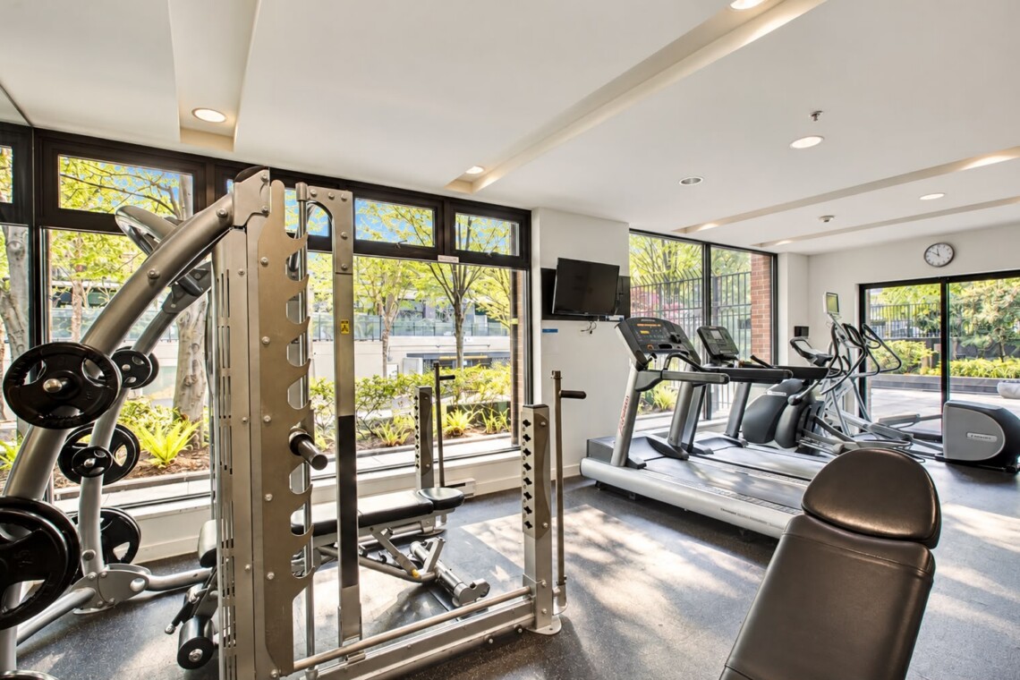 Well-lit gym with a multi-station weight machine in the foreground and treadmills by large windows overlooking trees outside. - Informative image with equipment layout.