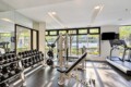 Bright gym interior with dumbbell racks, a multi-station weight machine, a bench, and cardio treadmills by large windows with greenery outside.