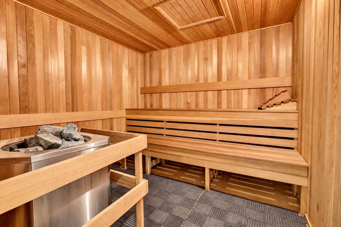Wood-paneled sauna interior with two-tier benches and a metal heater filled with rocks on the left.