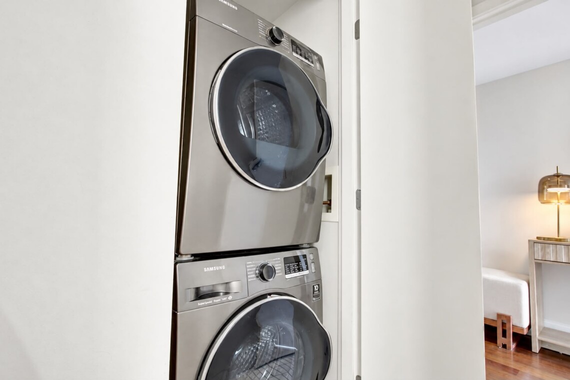 Stacked stainless steel Samsung front-loading washer and dryer in a laundry closet, with a living area and lamp visible to the right.