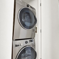 Stacked stainless steel Samsung front-loading washer and dryer in a laundry closet, with a living area and lamp visible to the right.