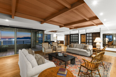Open living room with a wood beam ceiling, large glass doors to a balcony, and a view of water at sunset behind beige sofas and chairs:**Note: adjust for context**