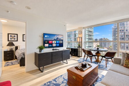 Open-concept living area with a wall-mounted TV and a dark wood credenza, plants, and large windows overlooking city buildings.