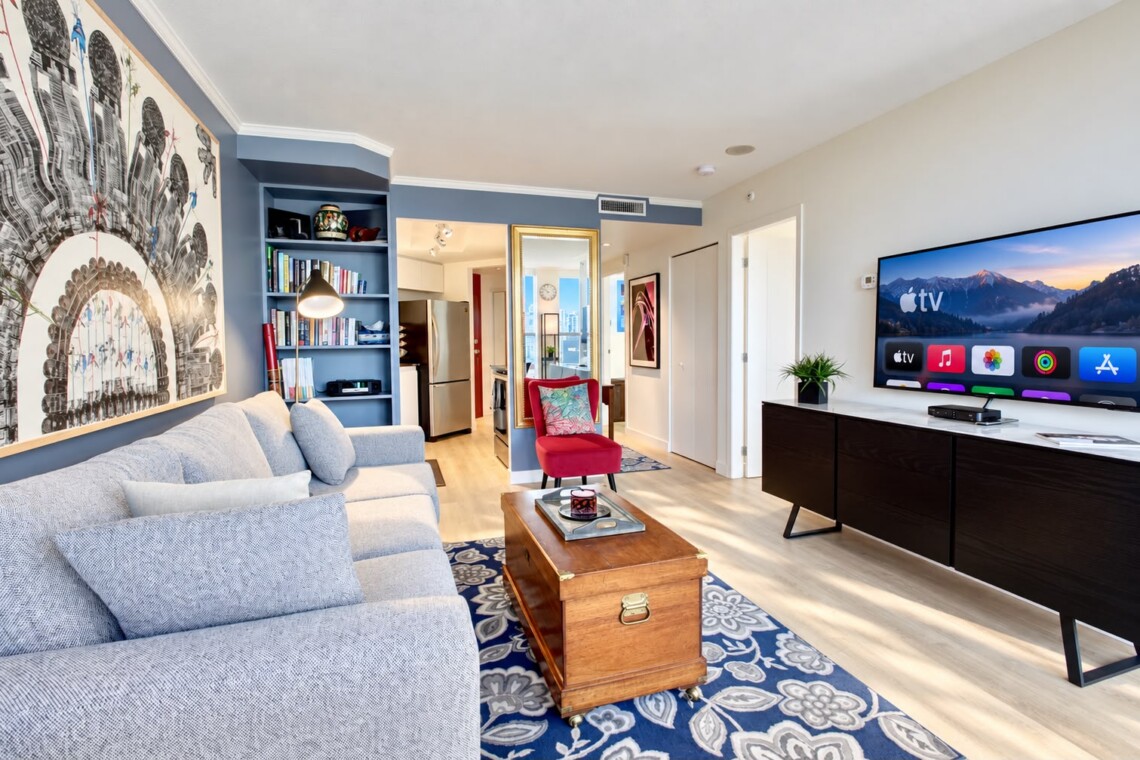 Living room with a gray sectional, blue accent wall, and a large architectural mural; open kitchen visible in the background.