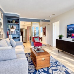 Living room with a gray sectional, blue accent wall, and a large architectural mural; open kitchen visible in the background.