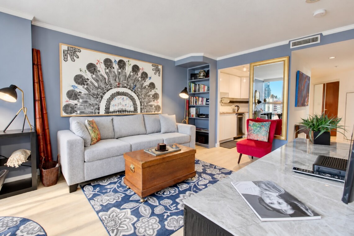 Living room with blue walls, a light gray sofa, and a large black-and-white semi-circular artwork above it; a wooden trunk serves as a coffee table on a blue floral rug, with a red chair nearby and a bookshelf nook to the side.