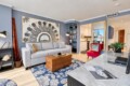 Living room with blue walls, a light gray sofa, and a large black-and-white semi-circular artwork above it; a wooden trunk serves as a coffee table on a blue floral rug, with a red chair nearby and a bookshelf nook to the side.