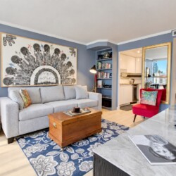 Living room with blue walls, a light gray sofa, and a large black-and-white semi-circular artwork above it; a wooden trunk serves as a coffee table on a blue floral rug, with a red chair nearby and a bookshelf nook to the side.