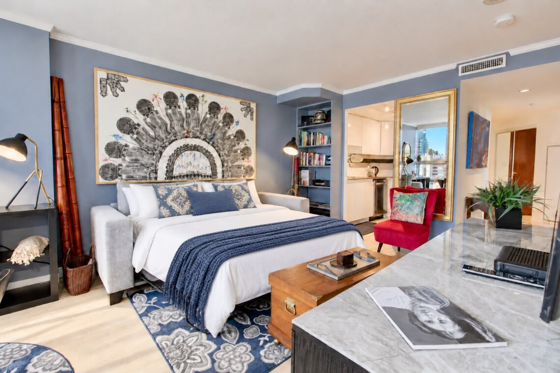 Modern hotel studio with a king bed, blue walls, large abstract artwork, sofa, and a marble-topped counter.
