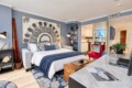 Modern hotel studio with a king bed, blue walls, large abstract artwork, sofa, and a marble-topped counter.