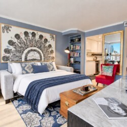 Modern hotel studio with a king bed, blue walls, large abstract artwork, sofa, and a marble-topped counter.