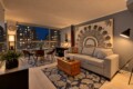 Cozy modern living room with a grey sofa, large artwork, and city lights visible through floor-to-ceiling windows at dusk.