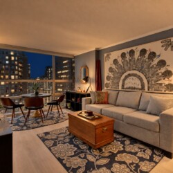 Cozy modern living room with a grey sofa, large artwork, and city lights visible through floor-to-ceiling windows at dusk.