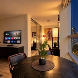 Open-concept living area at dusk with a round black dining table and a potted plant, a wall-mounted TV, and warm ambient lighting. Outside, city lights glow through a large window.