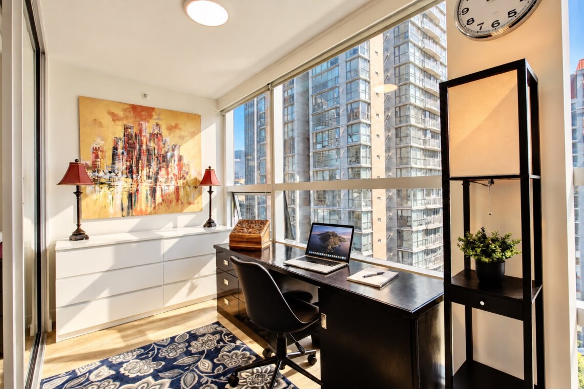 Bright corner home office with a black desk, laptop, and chair by expansive windows overlooking city high-rises; artwork, two red lamps, and a patterned blue rug nearby.