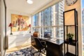 Bright corner home office with a black desk, laptop, and chair by expansive windows overlooking city high-rises; artwork, two red lamps, and a patterned blue rug nearby.