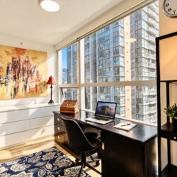 Bright corner home office with a black desk, laptop, and chair by expansive windows overlooking city high-rises; artwork, two red lamps, and a patterned blue rug nearby.
