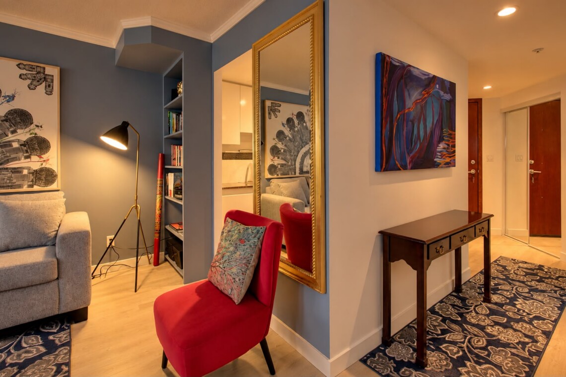 Cozy contemporary living area and hallway: blue accent wall, gray sofa, red accent chair with patterned cushion, tall floor lamp, built-in bookshelf, and a gold-framed mirror reflecting the space.