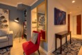 Cozy contemporary living area and hallway: blue accent wall, gray sofa, red accent chair with patterned cushion, tall floor lamp, built-in bookshelf, and a gold-framed mirror reflecting the space.