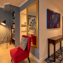 Cozy contemporary living area and hallway: blue accent wall, gray sofa, red accent chair with patterned cushion, tall floor lamp, built-in bookshelf, and a gold-framed mirror reflecting the space.