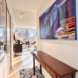 Narrow hallway opening to a bright living area with a large abstract painting on the right and a wooden console table with brass handles.