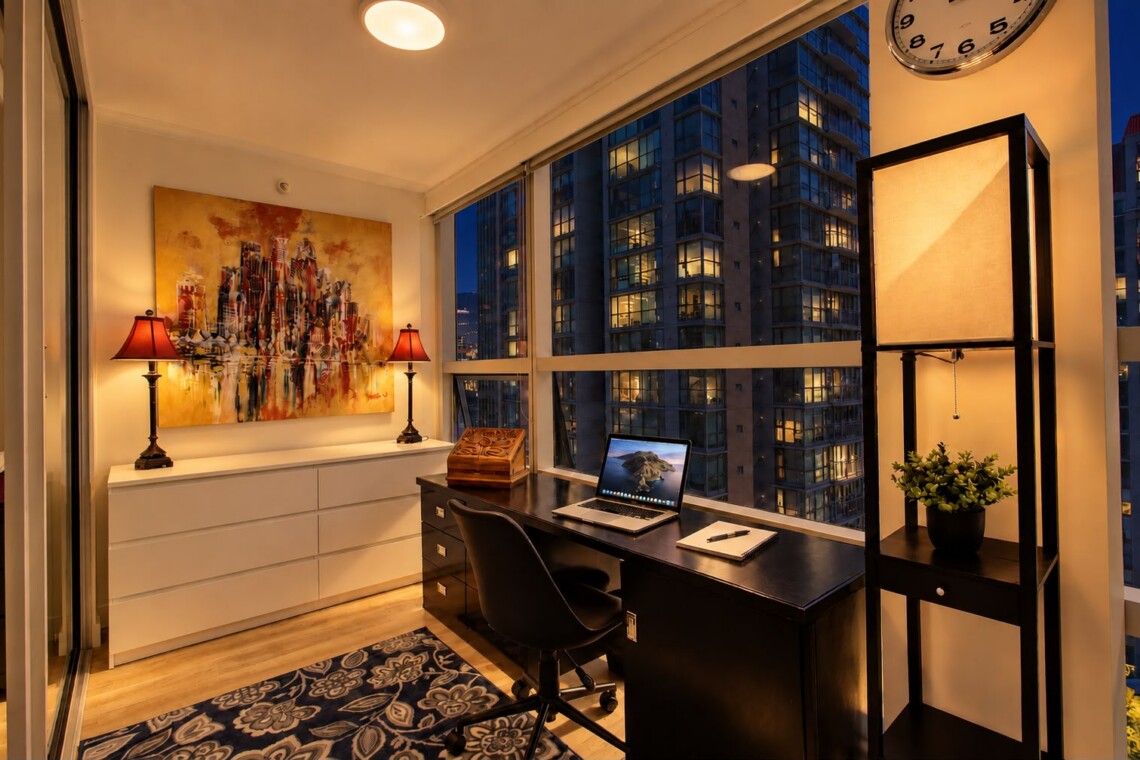 Modern home office in a high-rise apartment: black desk with laptop, notebook, and chair by large windows at dusk, with abstract painting and two lamps behind.
